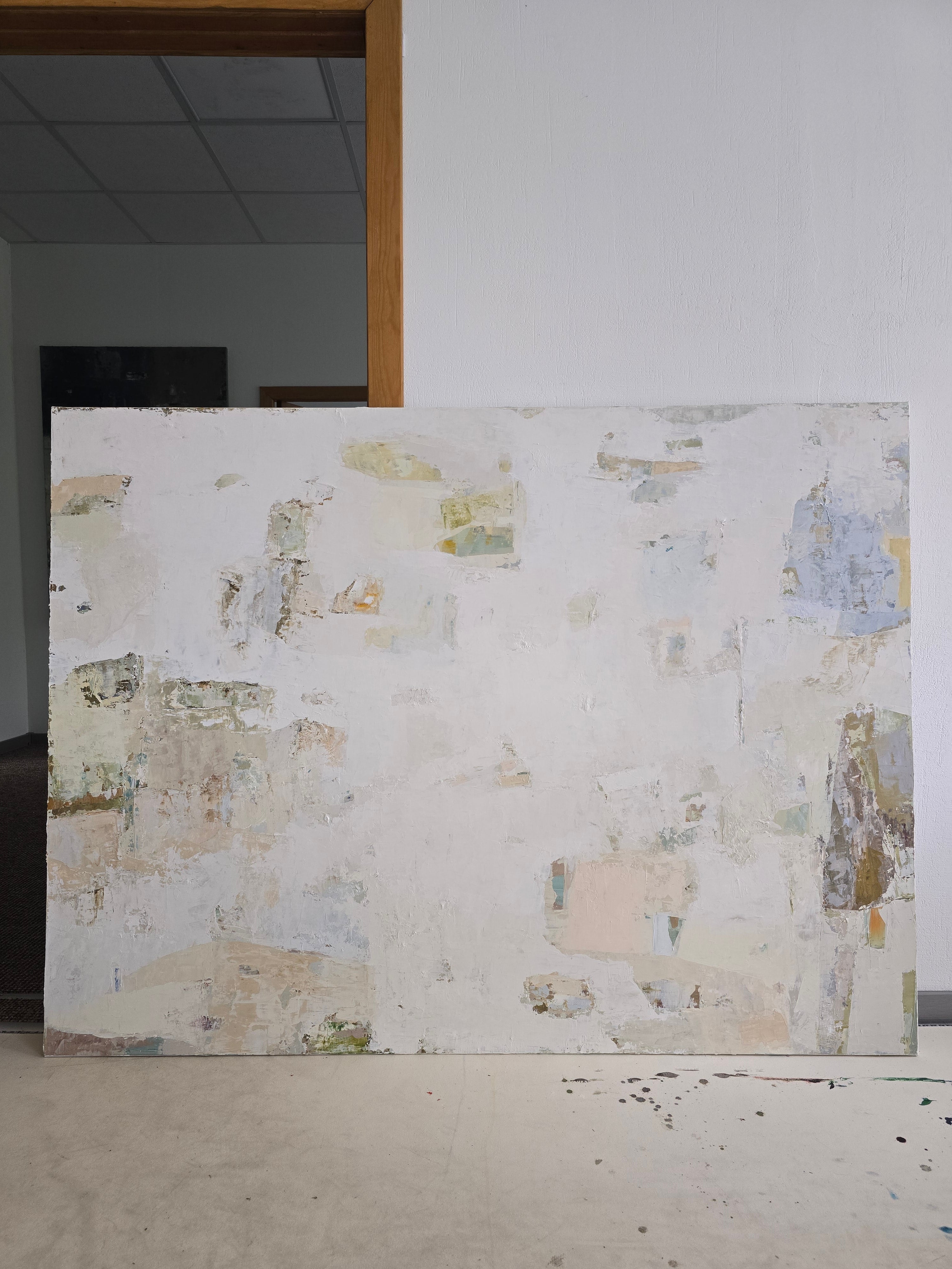 A large abstract  original canvas painting with a textured off-white background and patches of beige, light blue, and muted green, leaning against a white wall in an indoor space created by BIRUTE STUDIO.