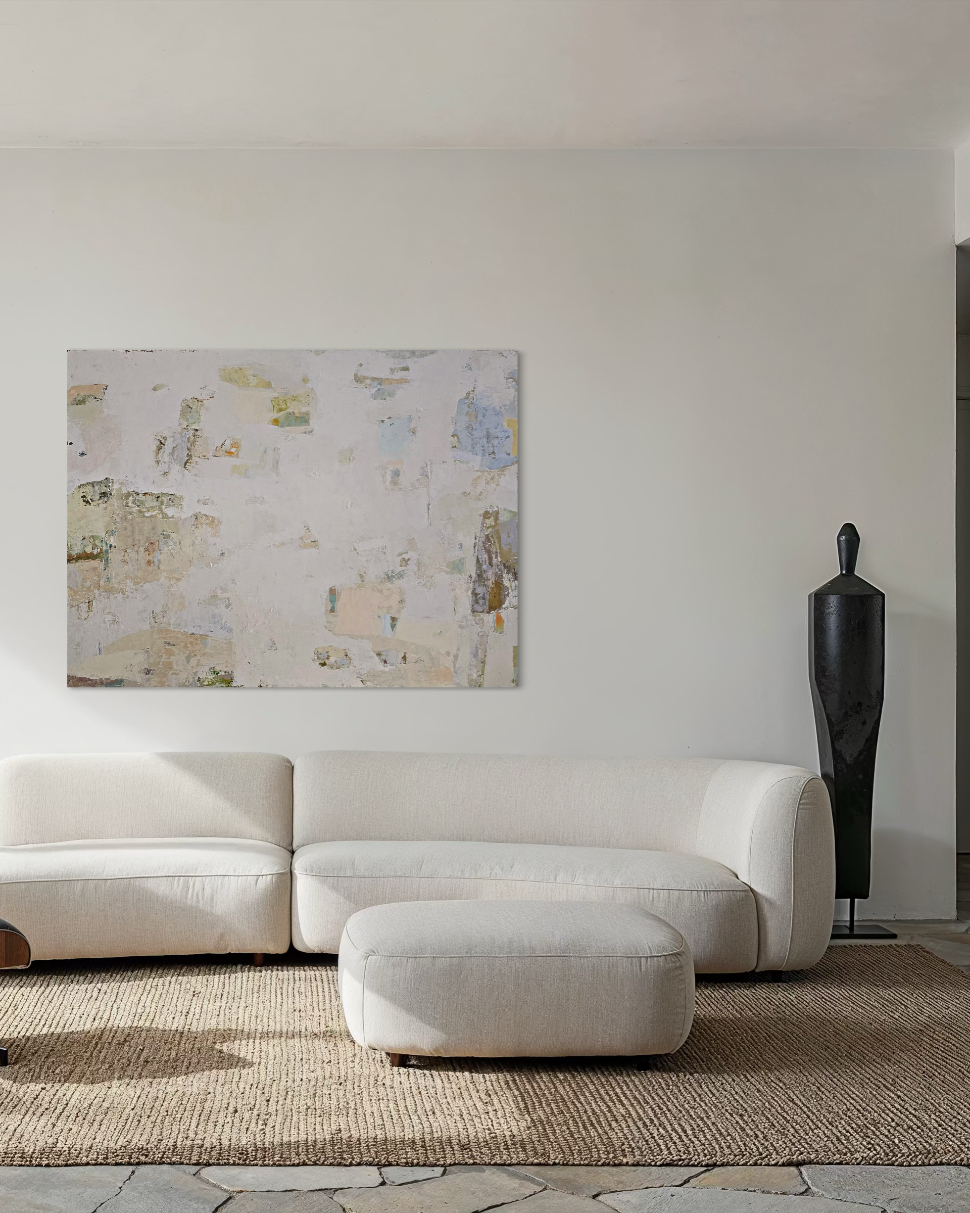 A modern living room scene featuring a cream-colored sectional sofa and matching ottoman on a woven jute rug, with a large abstract canvas neutral-tones painting on the wall.