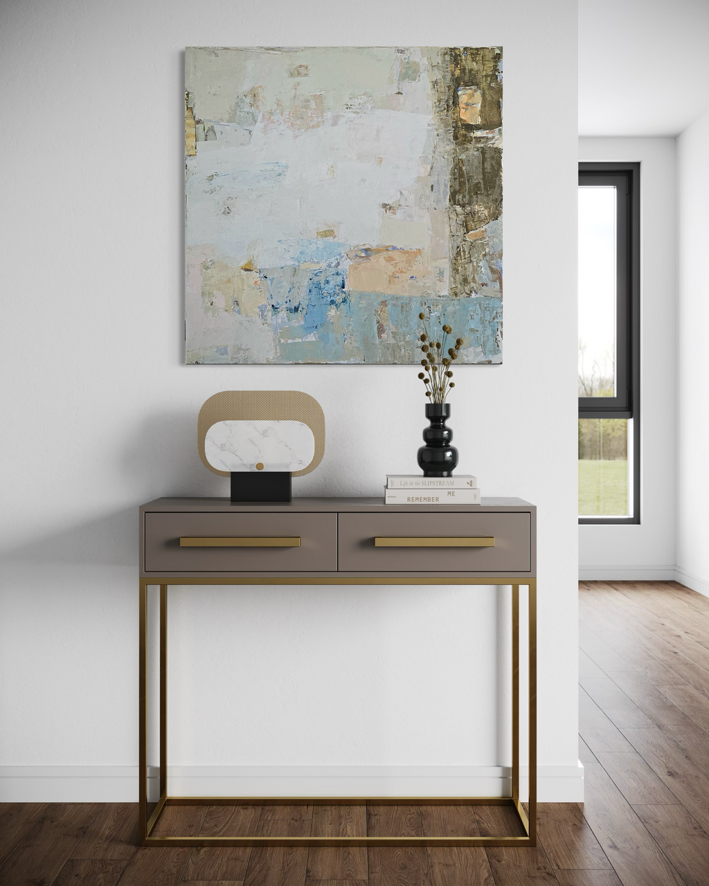 A modern abstract canvas painting in soft blue, beige, and neutral tones, displayed on a white wall above a gray console table with gold legs and handles in a bright room with wood floors.