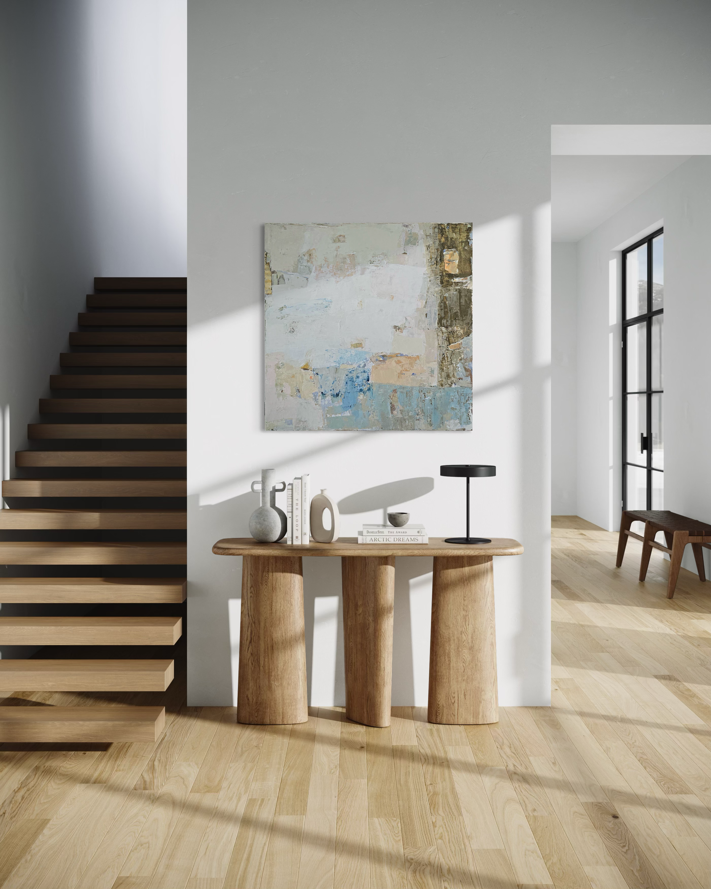 An abstract canvas painting with blue, neutral, and white tones hangs on a white wall above a wooden console table in a modern interior with a staircase and light wood flooring.