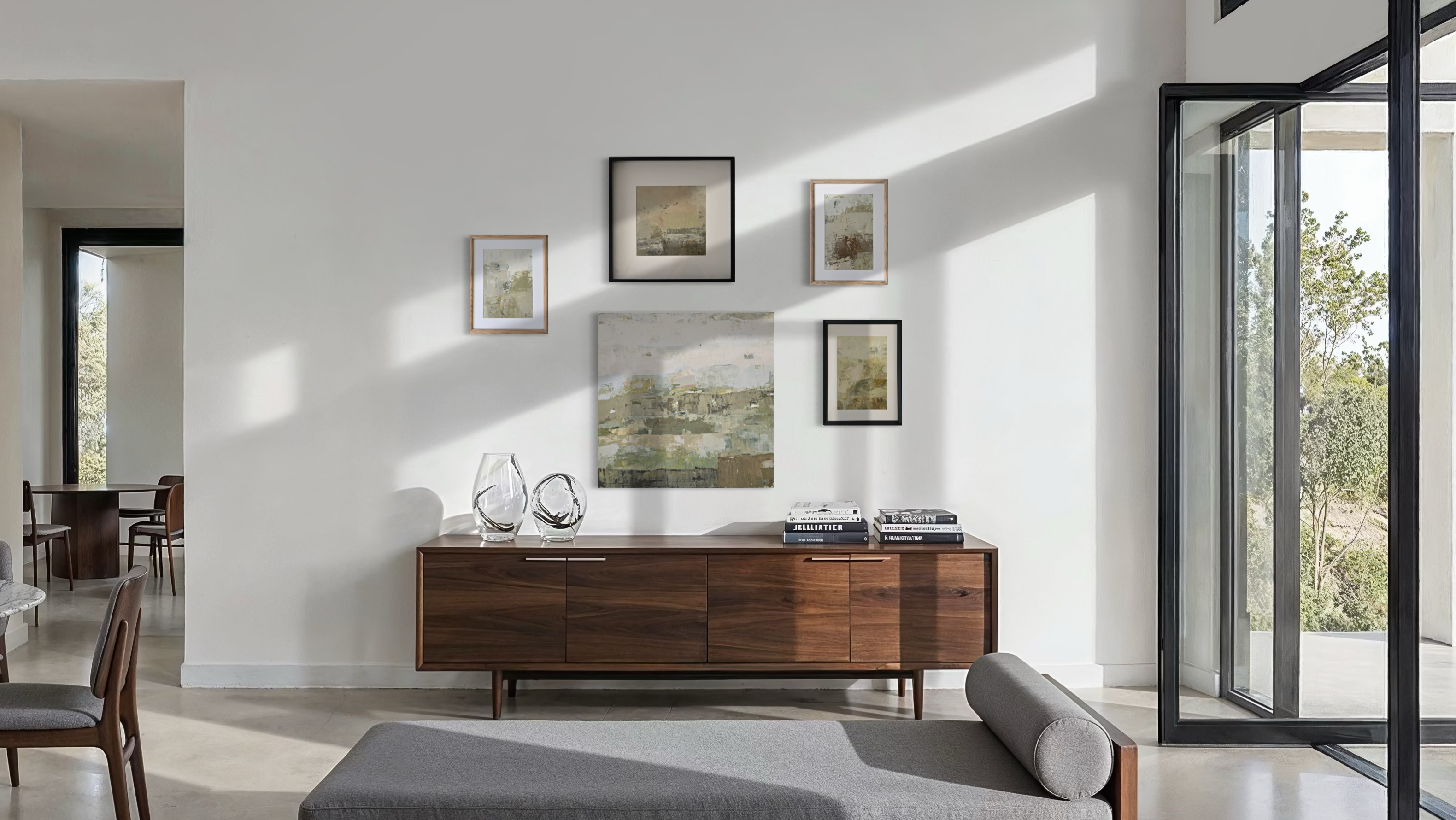 A mid-century modern wooden sideboard cabinet styled in a bright, contemporary living room with minimalist framed original wall art created by Birute Studio, decorative glass vases, and books on top.