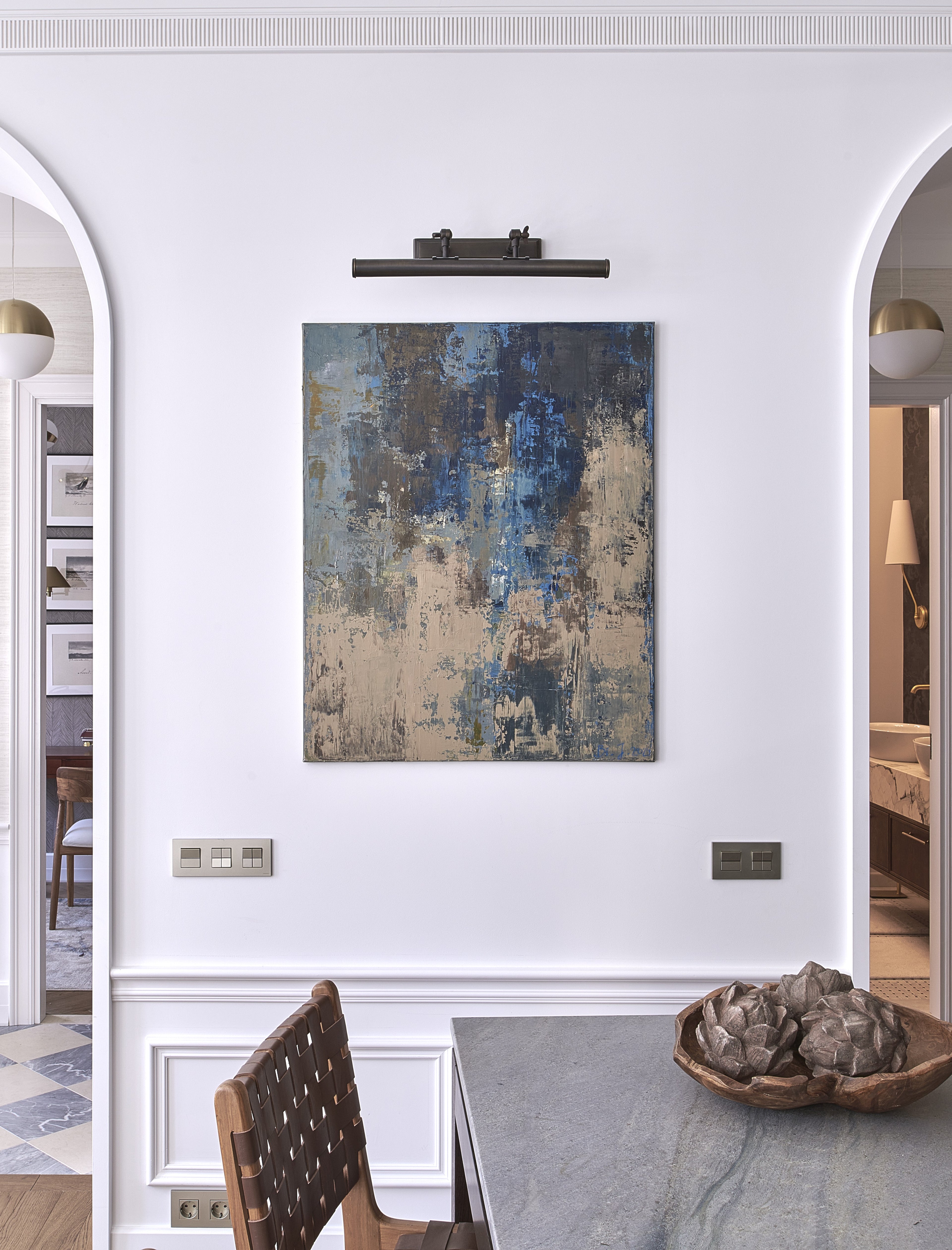 An abstract canvas painting with textured blue, gray, and beige created by Birute Studio and displayed on a white wall in a dining room setting above a gray stone table in Vilnius.